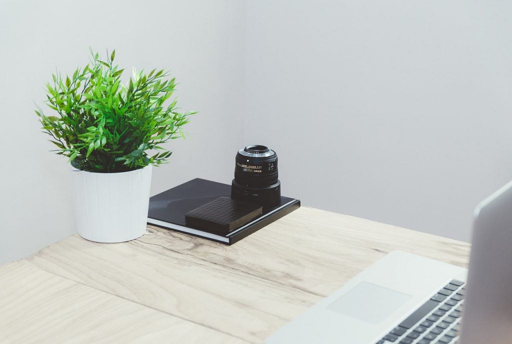 Auf einem Schreibtisch steht ein Laptop, ein Notizbuch, ein Kameraobjektiv und eine Grünpflanze