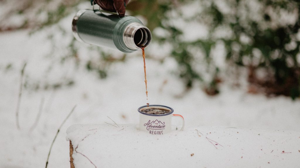 Von einer Thermoskanne wird Kaffee in eine im Schnee stehende Tasse gegossen
