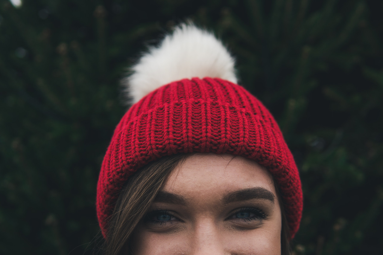 Vor einer Hecke steht eine braunhaarige Frau mit einer roten Bommelmütze auf dem Kopf